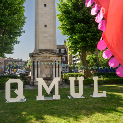 Large lettering spelling 'QMUL' on the green outside the Queen's Building