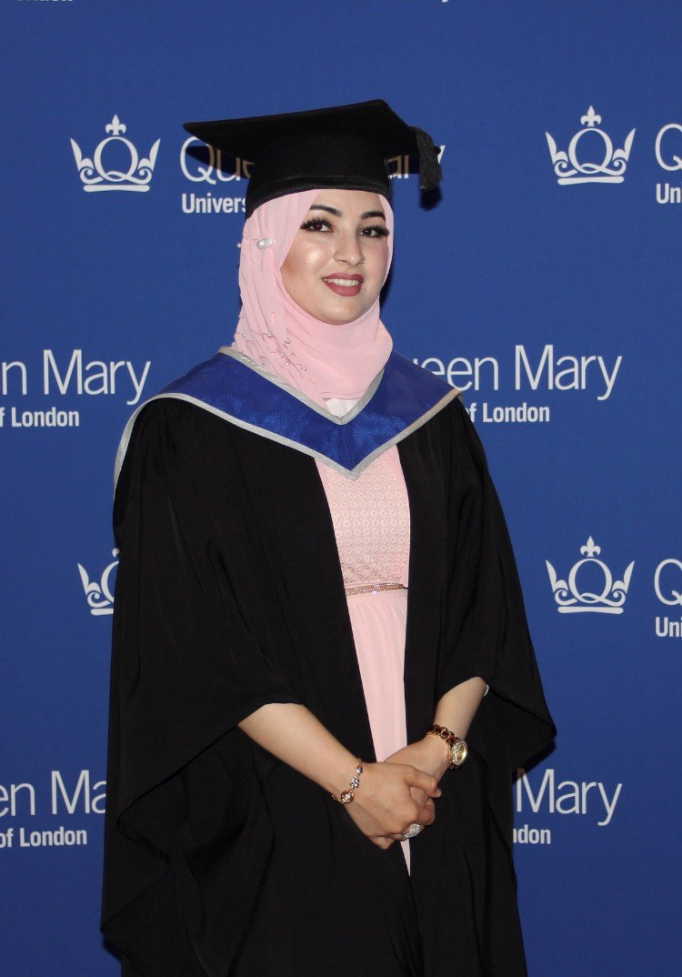 Amina in her graduation robes in front of a Queen Mary backdrop