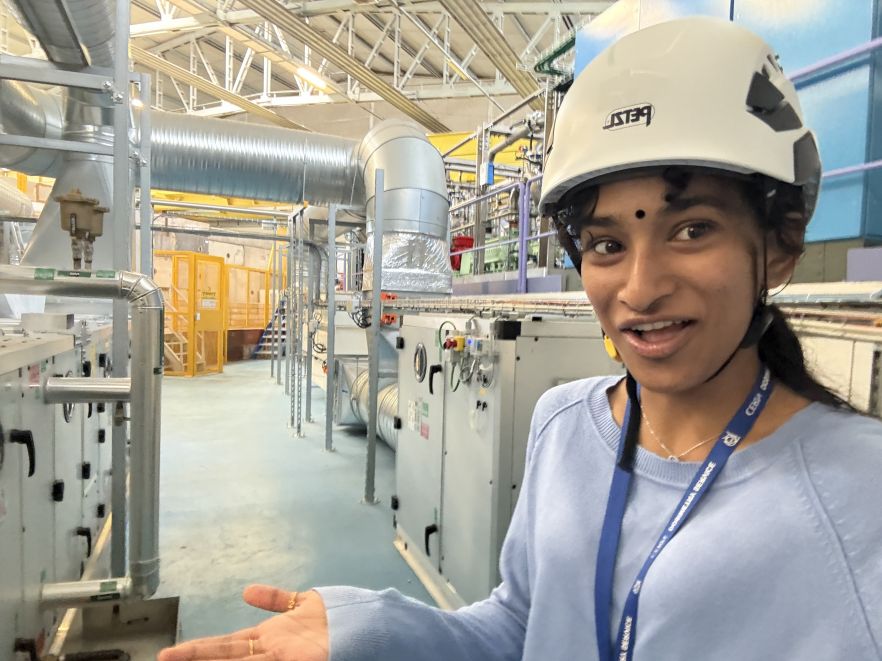Krishni in a hard hat, pulling a funny face as she gestures towards a large lab of technical equipment