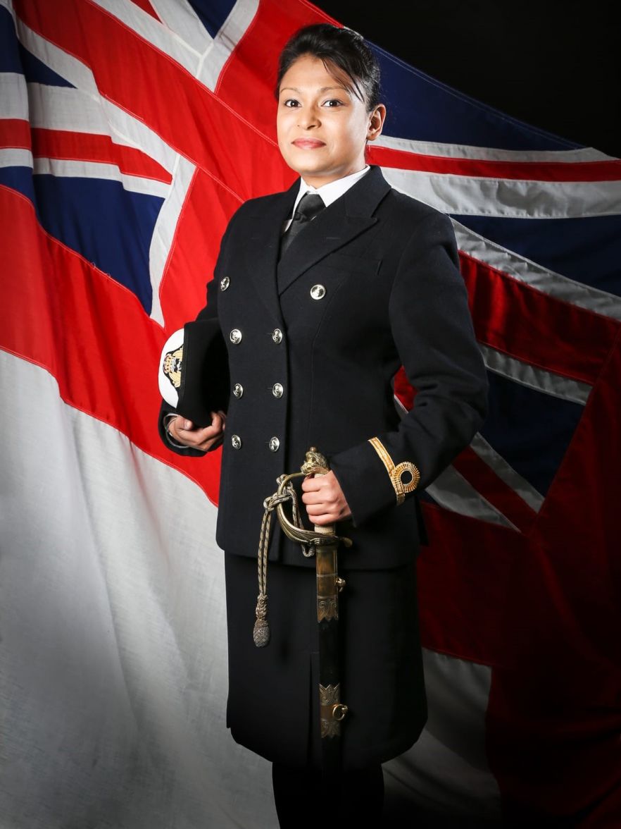 A woman wearing a military uniform standing proudly in front of a union jack, holding ceremonial sword and hat