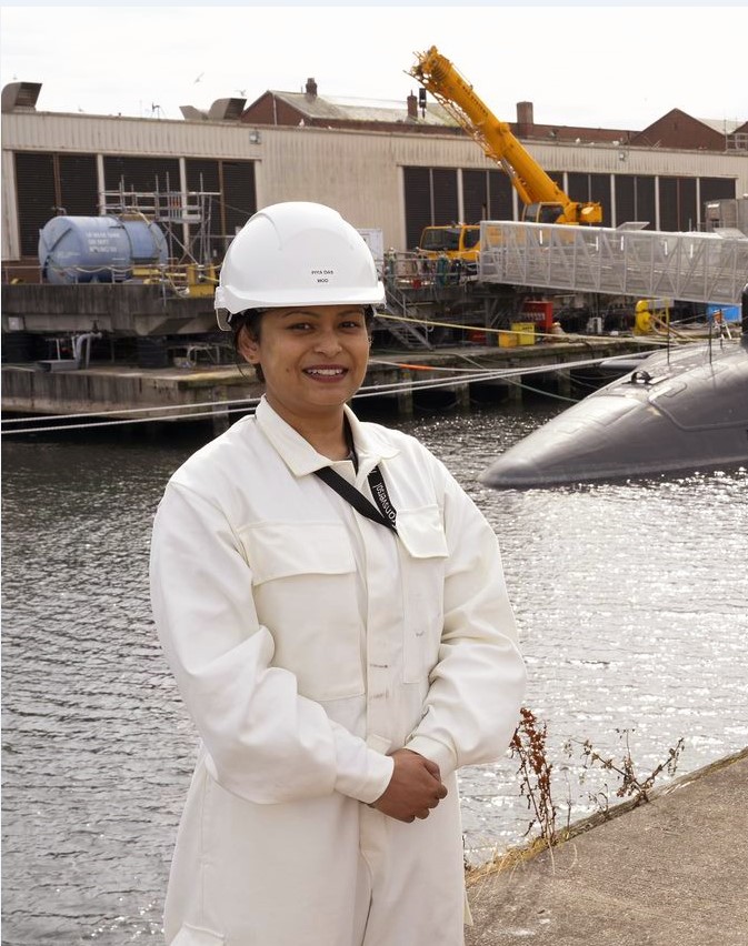 A woman in a white lab coat and hard hat in front of water with a submarine being built