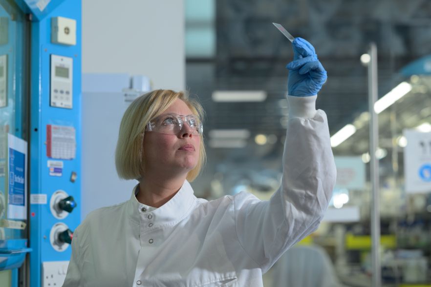 A scientist in a lab coat, goggles and gloves holding up and looking at something small
